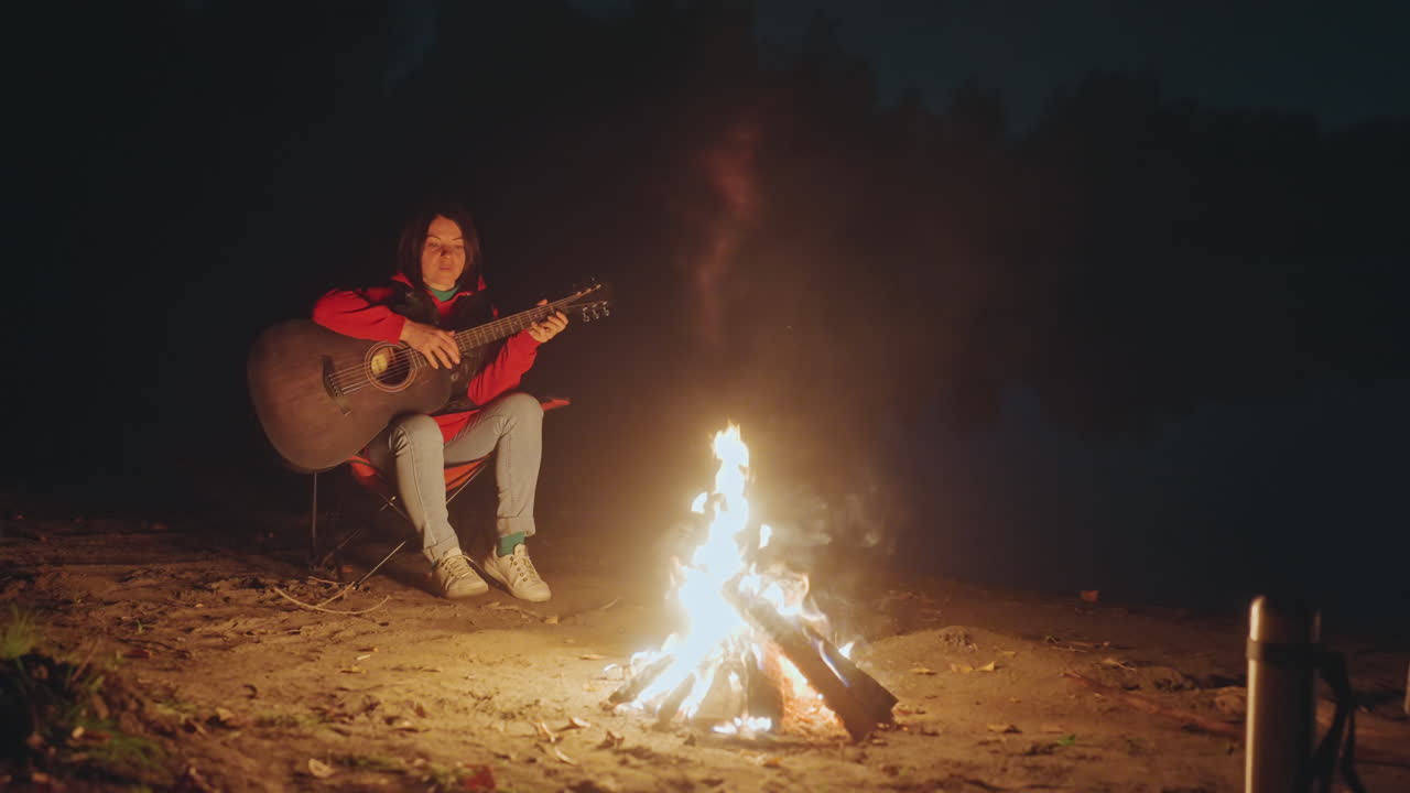 Noite à beira do lago, campista branco tocando violão, fogueira de acampamento aquecendo jaqueta jeans, sessão de composição acústica, fumaça flutuando sobre a areia, luzes distantes da cidade refletindo na água, músico solitário.