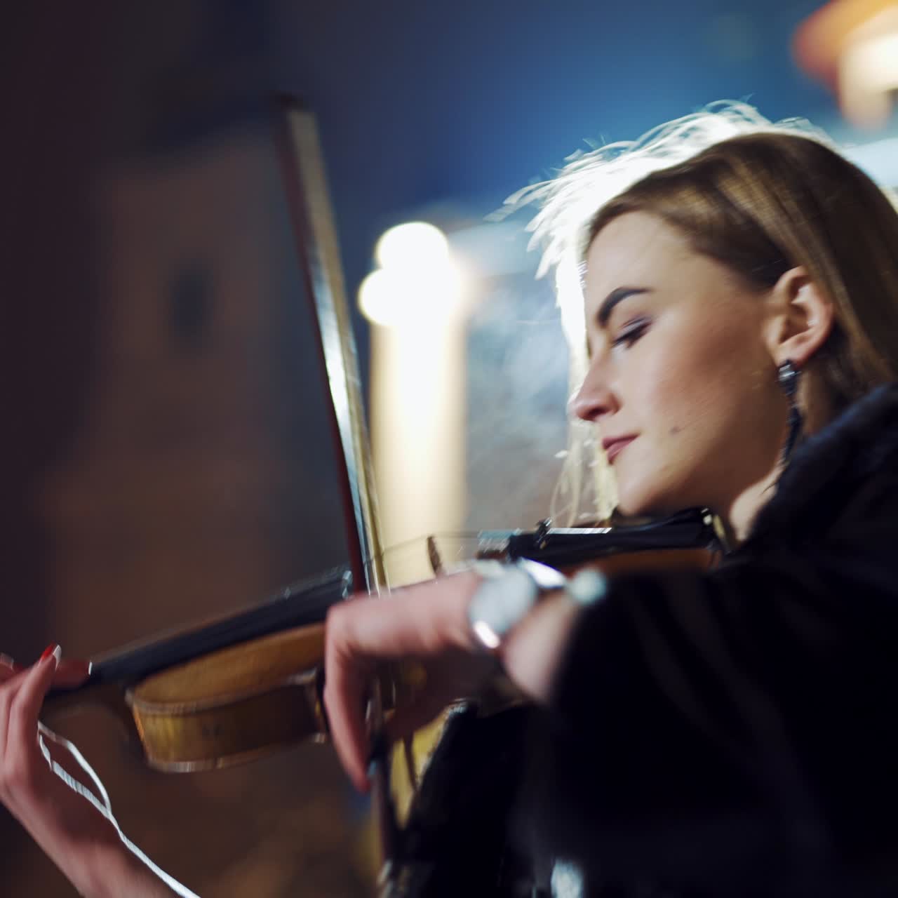 A charming girl with a watch on her hand is playing an instrument against the background of a large building in the evening in the winter. Violin. Close-up. Blurred background.