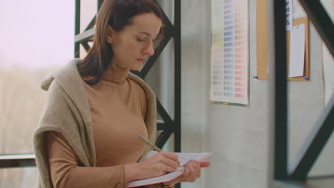 young woman colorist designer picks up colors standing near the wall. looking at the wall of sketches and flowers. choose the right color.