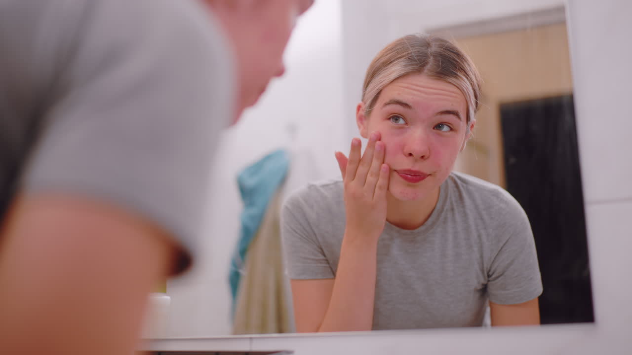 Independent young woman washing face over sink in bathroom, water splashing gently, reflection in mirror showing natural skincare routine, refreshing self care moment under soft indoor light