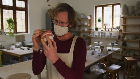 male caucasian potter wearing face mask and apron using brush to paint pot at pottery studio