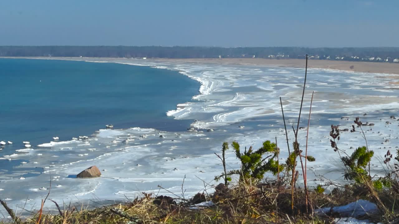 Sandy beach coastline is covered with frozen water creating a beautiful ice pattern during clear sunny day. High angle view from coast cliff over shore with calm blue water and empty frozen beach