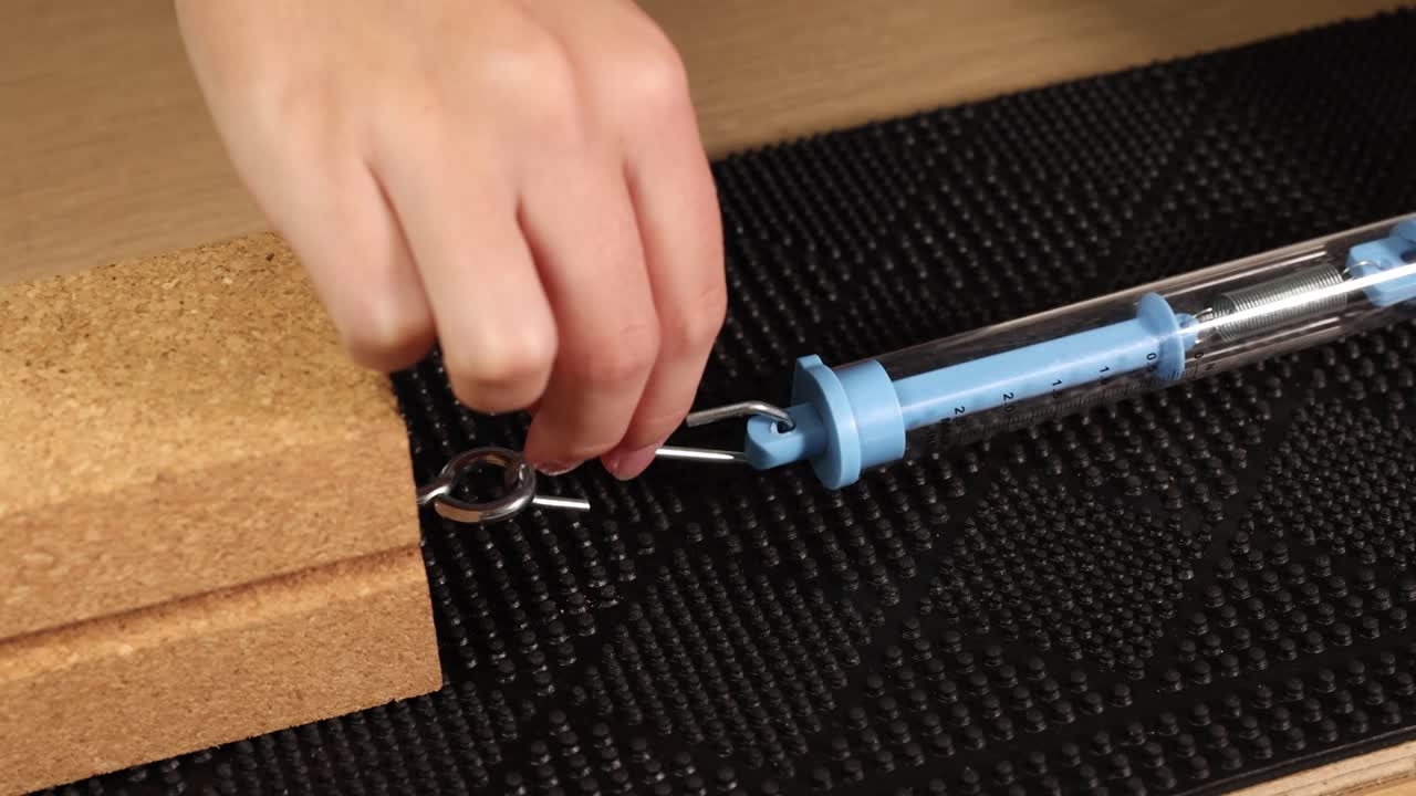 A hand uses a spring scale to measure frictional force on a wooden block across a textured surface under controlled lighting