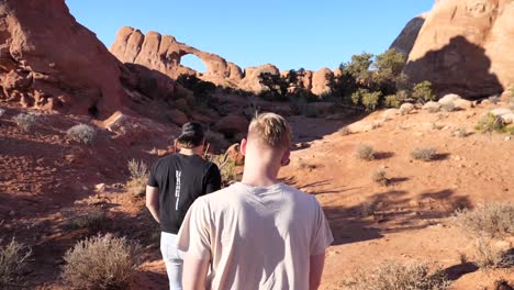 slo mo following two young men through red rock desert land with arch in foreground