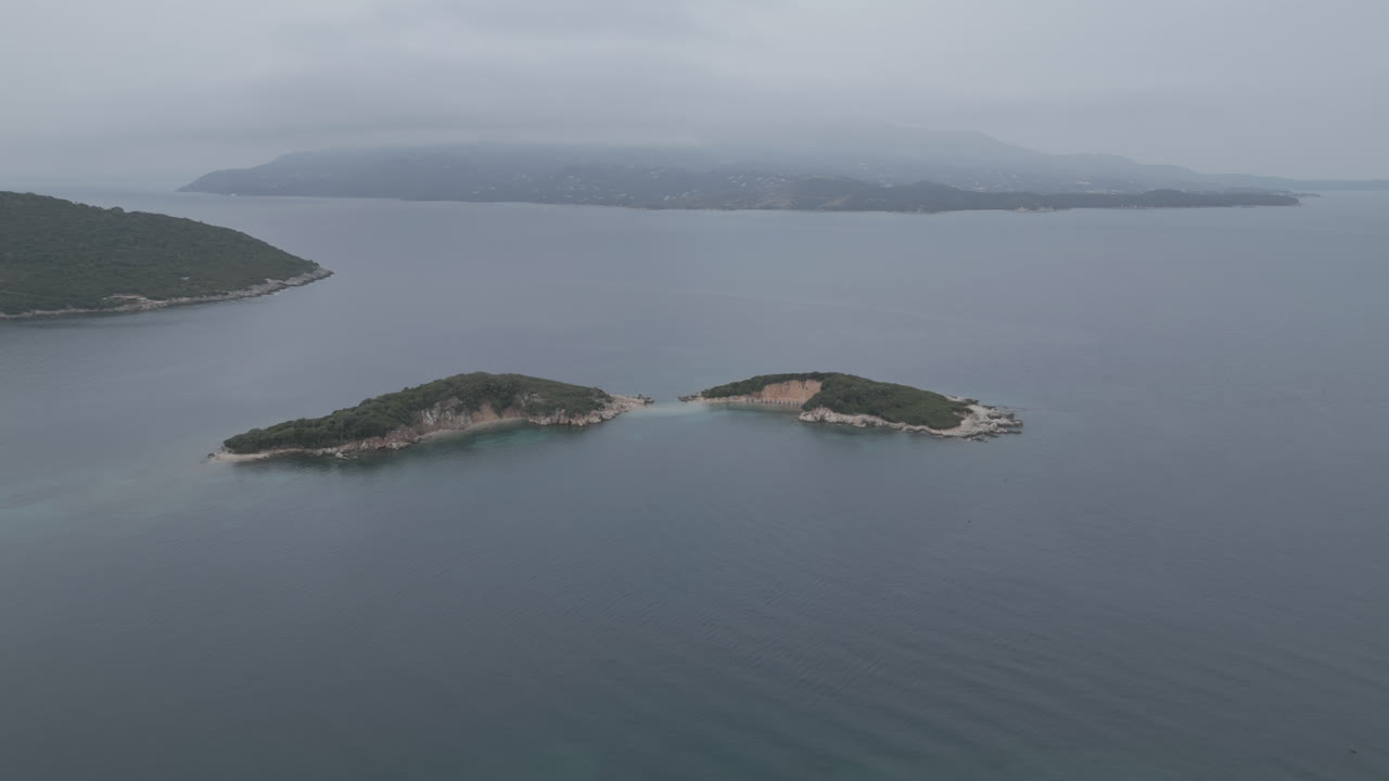 드론이 물 위를 날아서 크사밀 알바니아 앞의 작은 섬으로 날아가는 사진입니다. 구름이 많은 회색 날 아침에 배가 주변에 있고 하늘에는 안개가 있습니다.