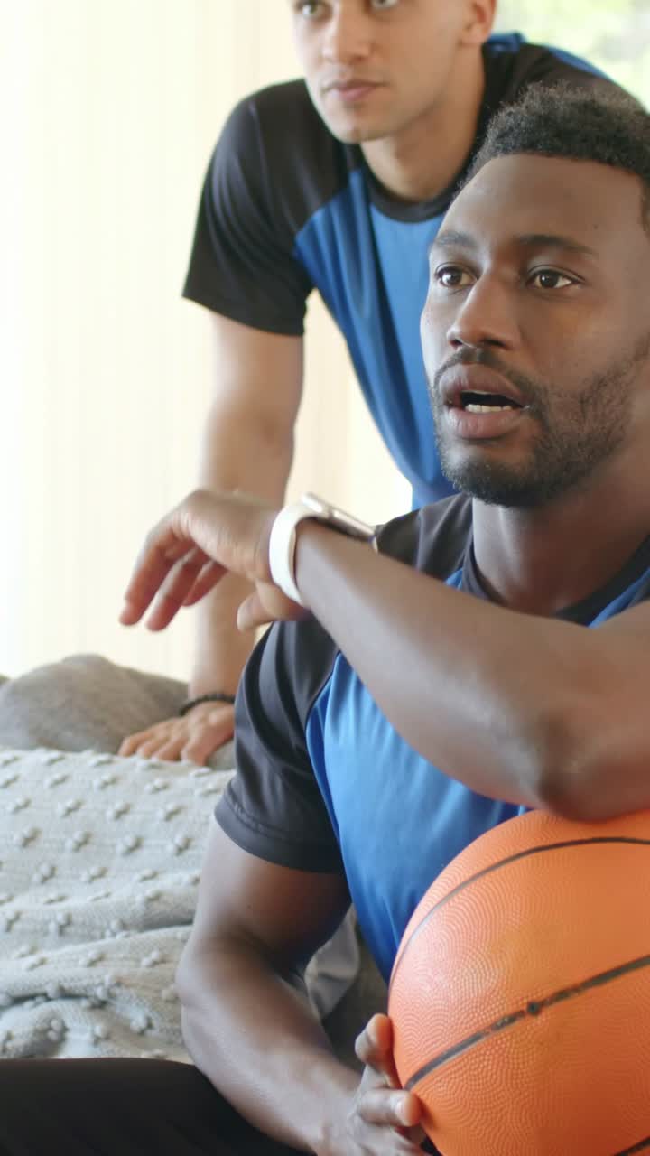 Vertikal video: Ser spil uden for skærmen, siddende mand i blå skjorte holder en basketball på sofaen derhjemme