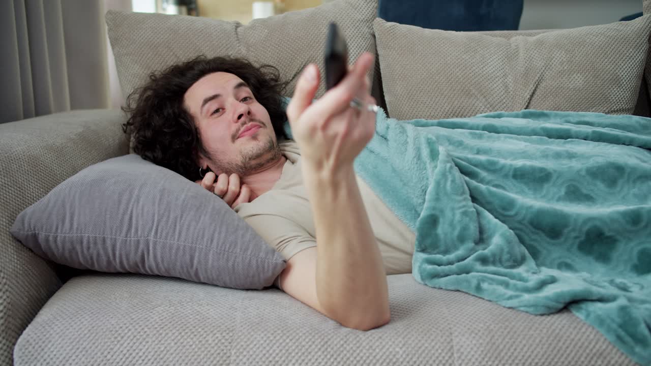 A happy brunette guy with curly hair and a mustache lies on a gray pillow with a blue blanket and switches channels using the remote control at home on the sofa on the weekend