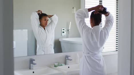 Mujer-Afroamericana-En-Albornoz-Atándose-El-Pelo-Mirándose-En-El-Espejo-Del-Baño