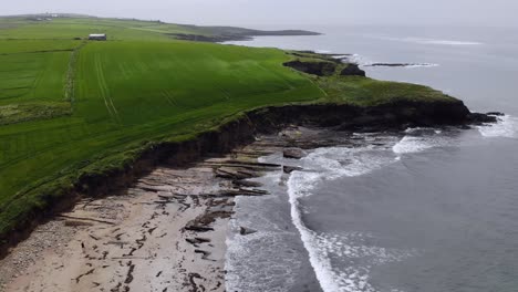aerial footage of a green farmland at the edge of the ocean with rugged cliffs and waves