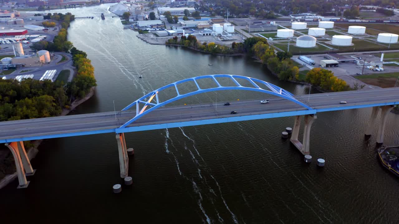 Drone captures a wide view of a blue arch bridge stretching across a calm river channel beneath a soft evening sky, showcasing structural symmetry and the blend of industry and nature