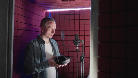 a man in a blue shirt and white inner shirt enters a red soundproof room, preparing to sing as he puts on headphones and stands ready at the microphone