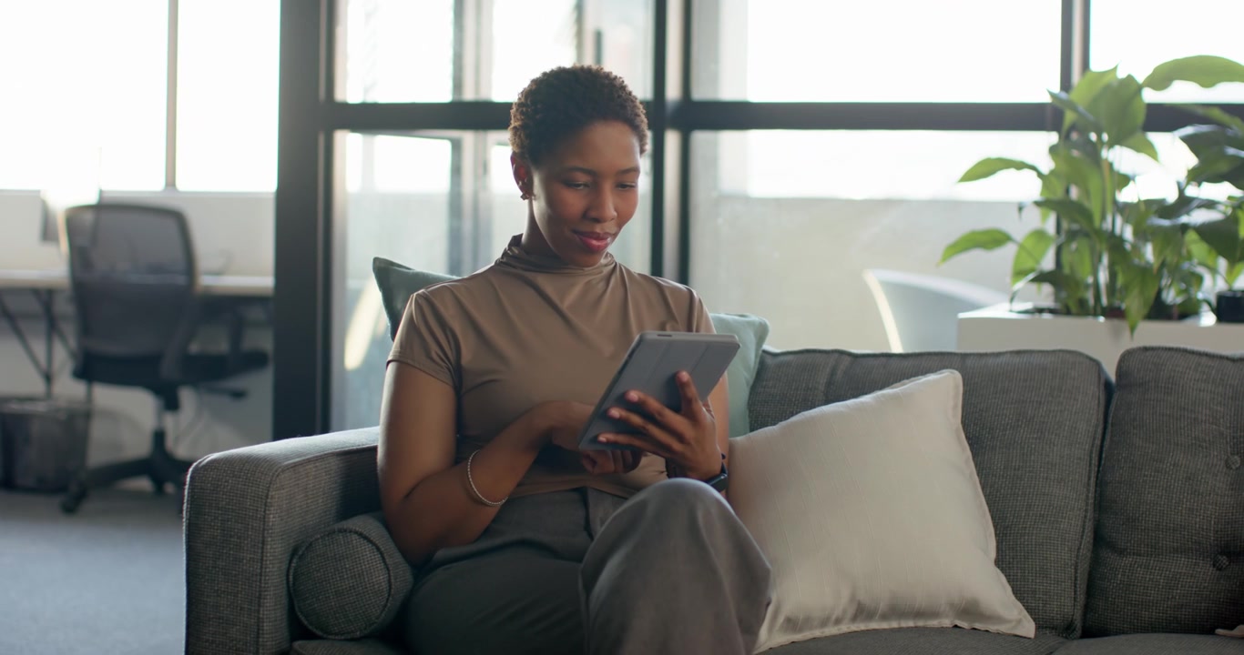 African woman settling on sofa in office wearing taupe top tapping, scrolling tablet reading emails