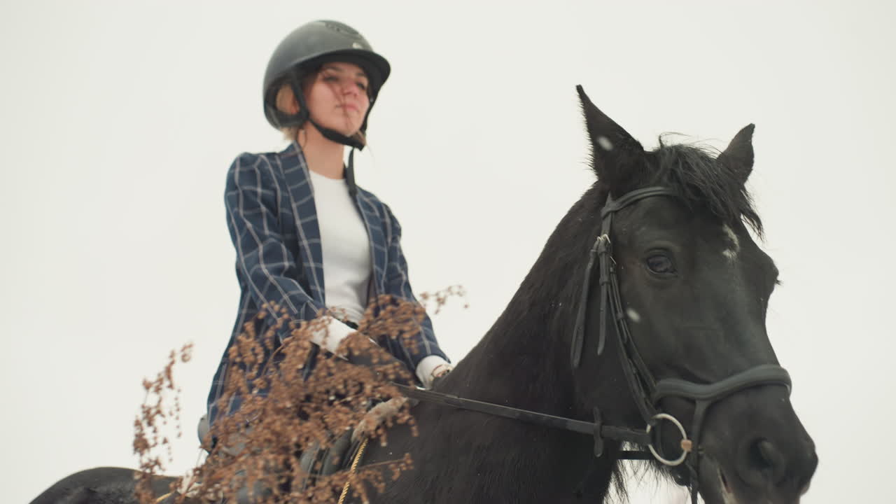 Donna bianca su cavallo nero in posa con casco e blazer, che pratica una lezione di equitazione in tandem, espressione calma, redini in mano, prato secco in primo piano, cielo coperto, primo piano di allenamento all'aperto