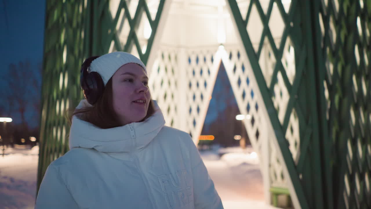 Young lady wearing white winter coat and headphones moves body cheerfully beside illuminated gazebo under night sky with snow covered ground and warm glow from overhead lights