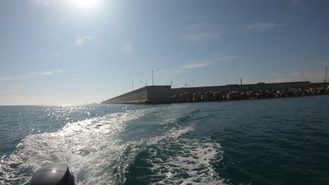 Retroiluminación-De-La-Popa-De-Un-Barco-Que-Se-Aleja-Del-Puerto-Con-El-Sol-En-La-Parte-Superior-Del-Avión-Y-Las-Paredes-Del-Puerto-A-Lo-Lejos,-La-Estela-Del-Barco,-El-Mar-Azul-Y-El-Cielo-Despejado