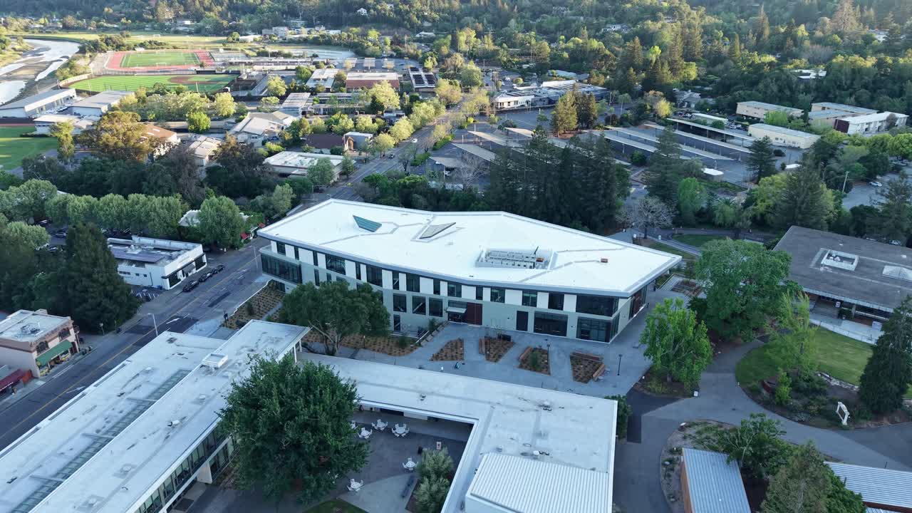 Vista aérea con dron alejándose lentamente sobre la arquitectura moderna del campus del College of Marin, rodeado de exuberantes árboles verdes y el paisaje urbano de Kentfield, California, al amanecer.