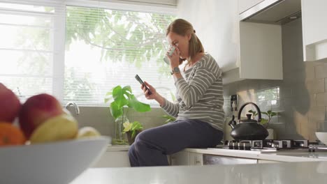 mujer caucásica bebiendo café y usando un teléfono inteligente en la cocina en casa