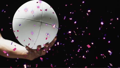 animation of confetti over white rugby ball held by caucasian man's hand on black background