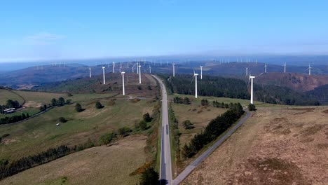 drone shot traveling forward on a road passing between a wind turbine park in the mountains, sunny day of blue sky with fog on the horizon