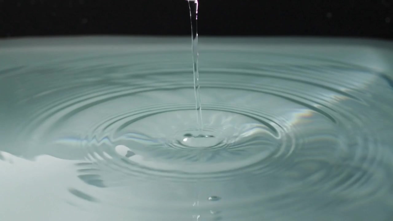 Eine weiße Düse gibt einen Wasserstrahl ab, der auf ein flaches Becken trifft und Wellen und Tropfen bildet.