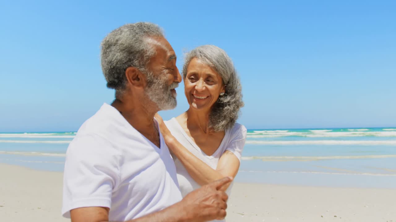Side view of happy active senior African American couple walking on beach in the sunshine 4k