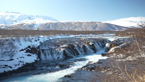 Beautiful Brúarfoss waterfall, Iceland, with turquoise water cascading over dark rocks. Snowy banks and mountains under a clear sky.
