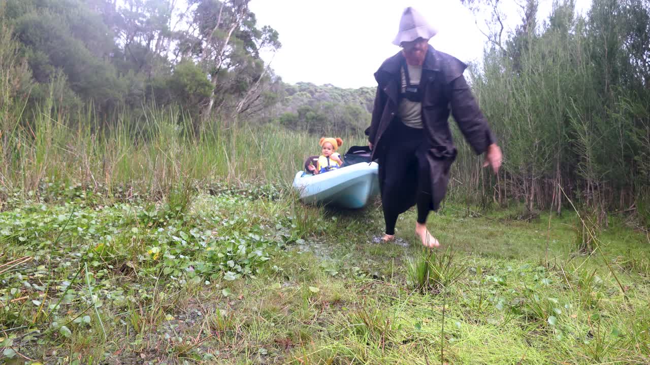 A man in a rain jacket drags a kayak through swamp land with his daughtet sitting in the kayak.
