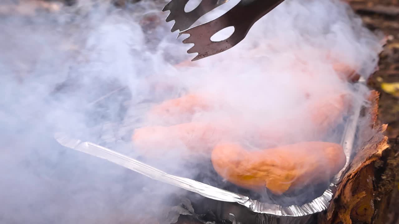 Nærbilde av pølser som grilles på en røykfylt engangsgrill, med grillklyper som snur dem over glødende kull utendørs