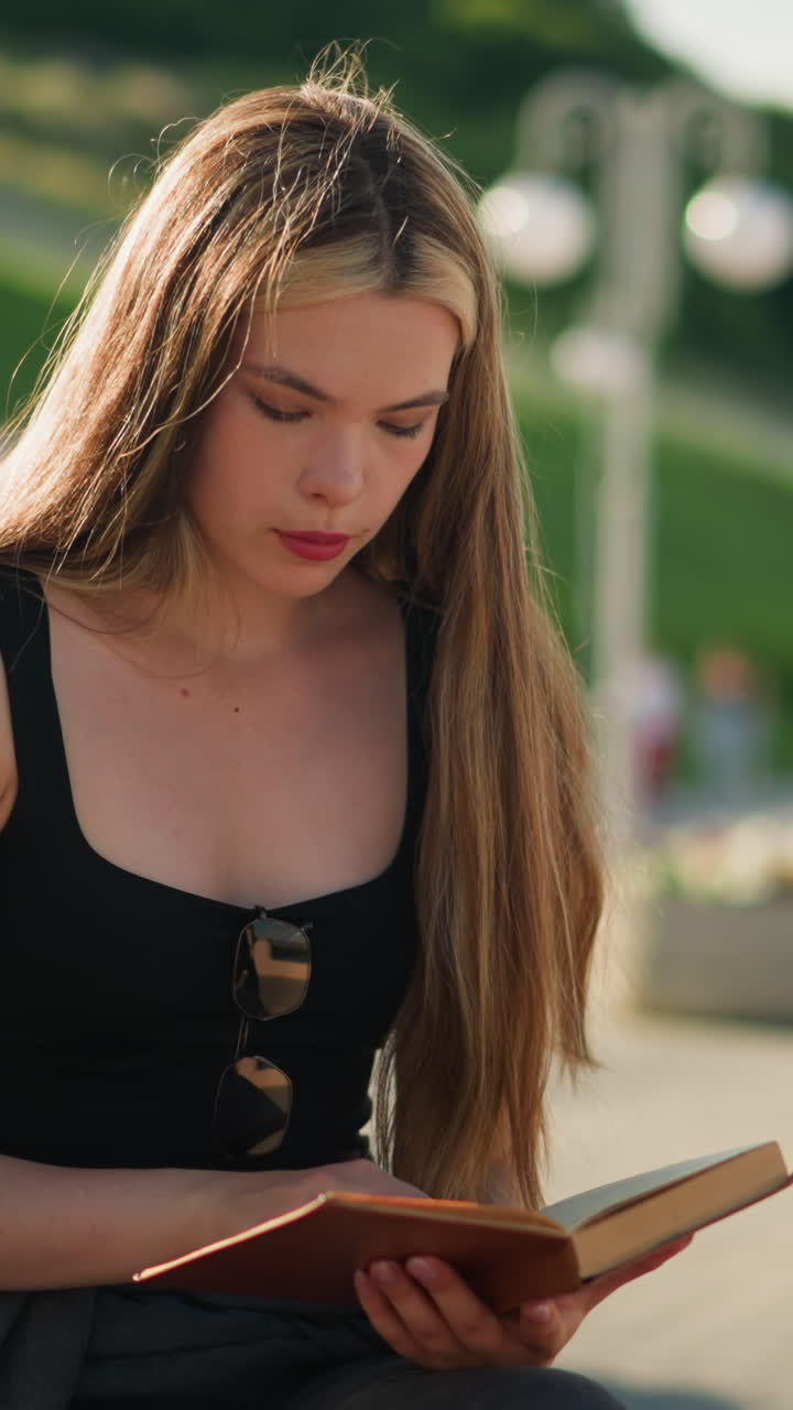 Woman seated outdoors on a bench with legs crossed, flipping a page from her book and thoughtfully observing a piece of paper, then placing it back, the background features blurred people walking