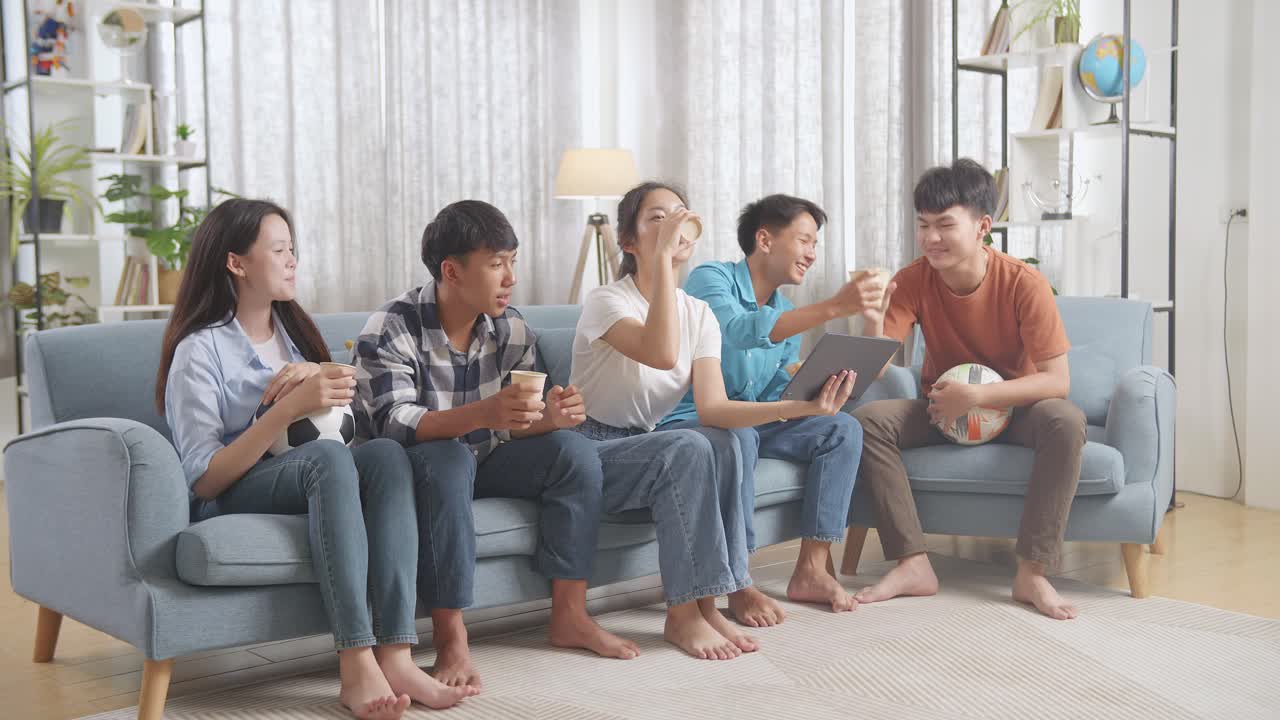 아시아 십대 들 이 집 에서 태블릿 에서 축구 경기 를 보는 동안 환호 하고, 먹고 마시고 있다