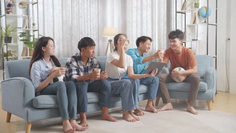 아시아 십대 들 이 집 에서 태블릿 에서 축구 경기 를 보는 동안 환호 하고, 먹고 마시고 있다