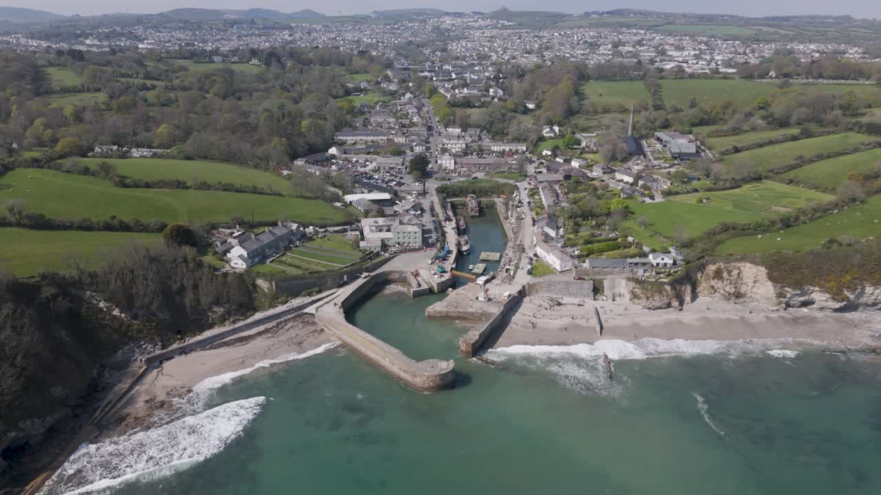 Aerial pan over picturesque seaside village with terraced homes lining a tidal harbor, narrow lanes leading to the beach and sea wall amid rolling rural hills