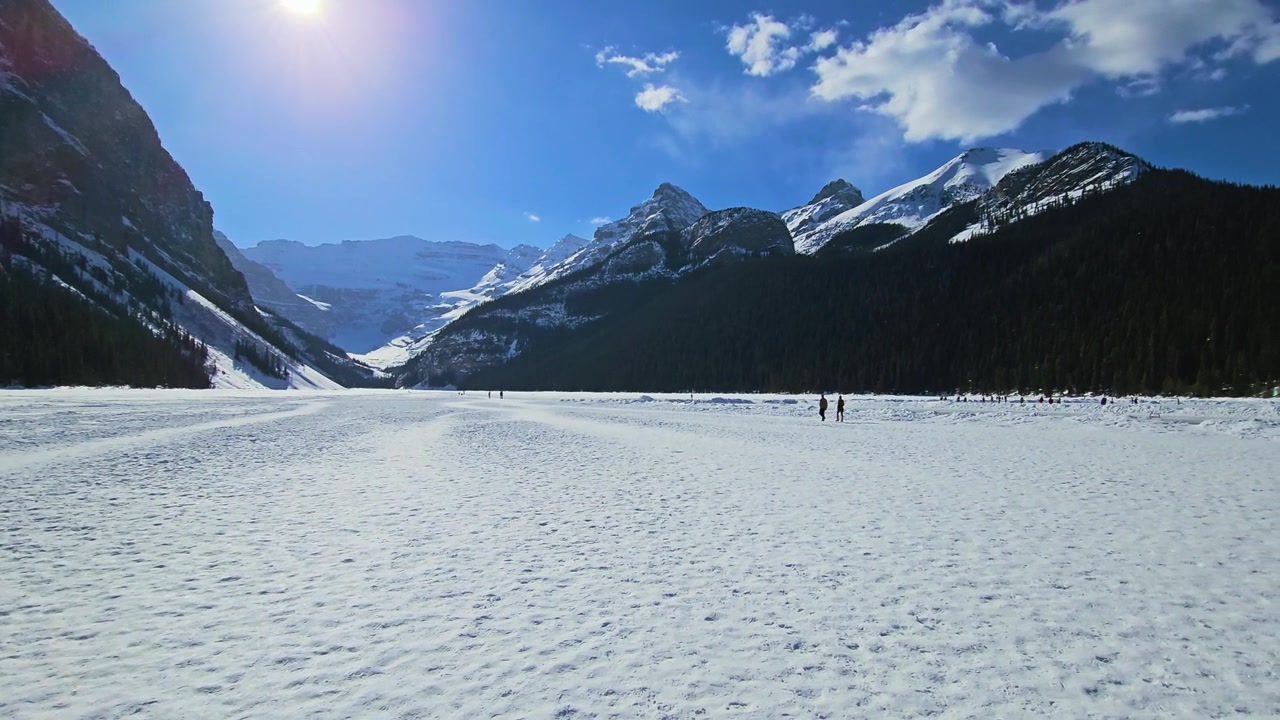 Persone camminano sul Lago Louise ghiacciato nel Parco Nazionale di Banff, circondate da montagne innevate, fitta foresta di conifere, luce diurna intensa e uno spettacolare paesaggio delle Montagne Rocciose canadesi