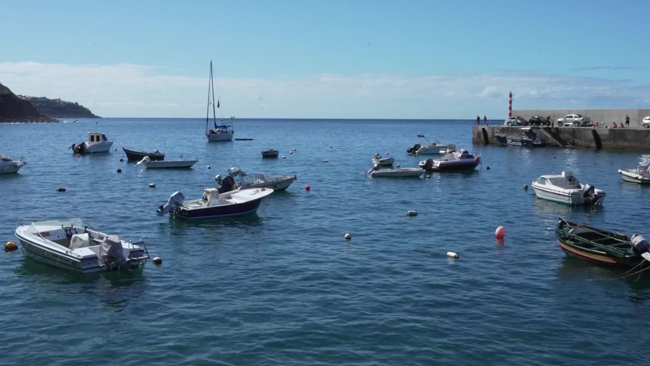 Riprese che mostrano il molo e le barche ormeggiate a Paul do Mar, incorniciate da ripide scogliere e dal vasto Oceano Atlantico. La scena enfatizza la scala e la bellezza naturale