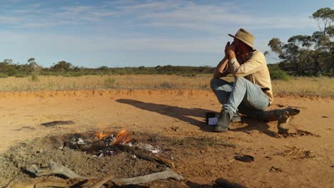 an authentic looking australian bushman play his harmonica by a campfire in the outback