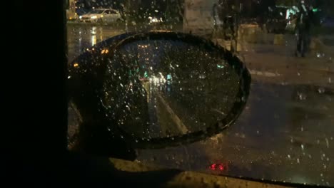 waiting in a car at a traffic light in the night on a rainy day. looking into a side mirror with cars passing by at night. rain drops on the car window. busy street at rainy night.