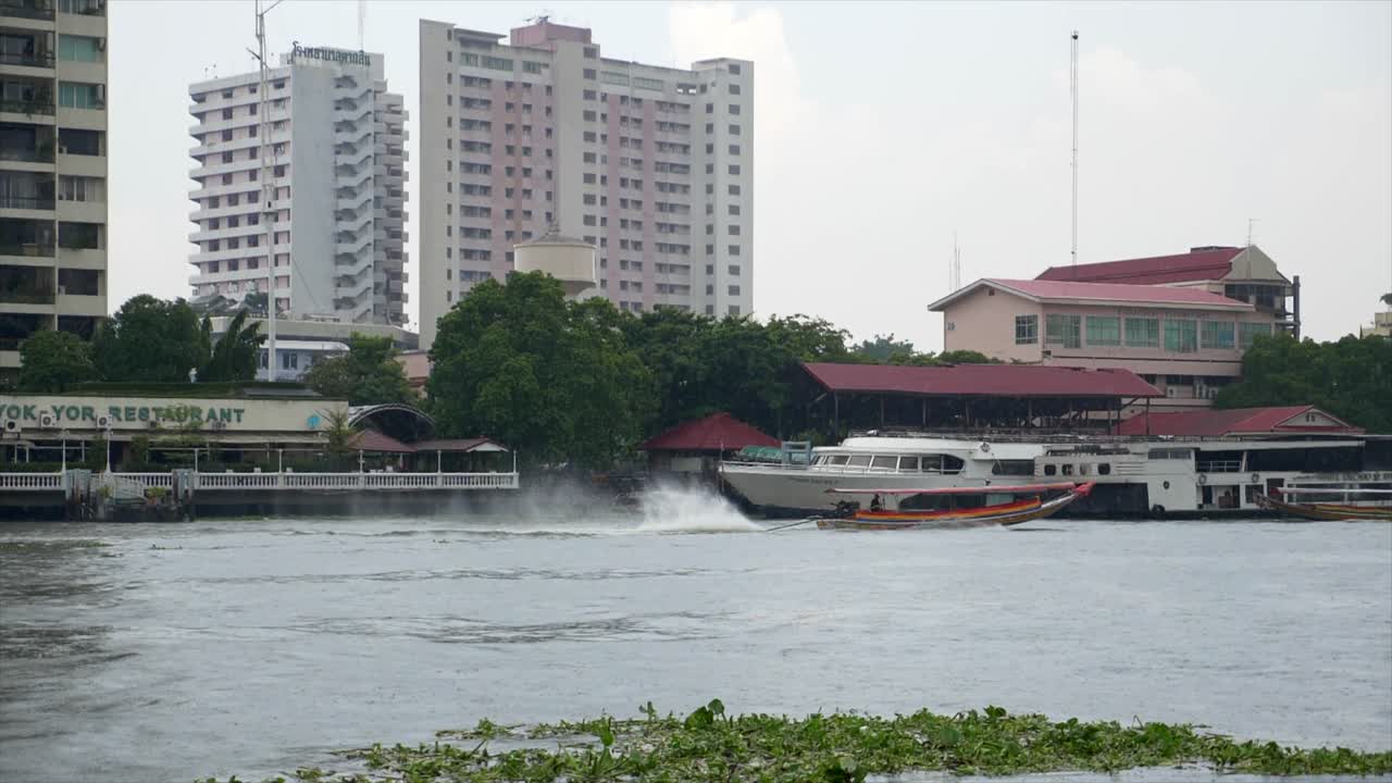 Slow motion shot following a motor boat sealing at high speed over the water of the Chao Phraya river in Bangkok over a background of traditional building and skyscrapers.