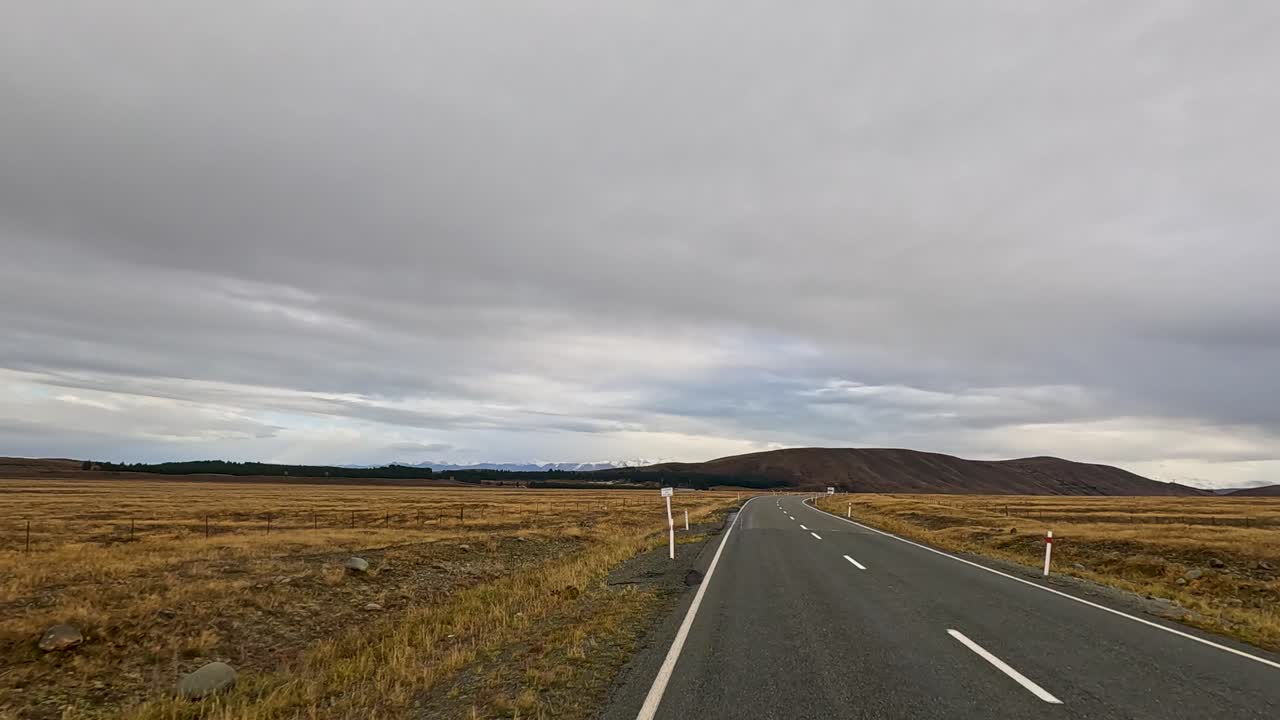 A white campervan travels along an empty rural road through golden autumn fields under overcast skies near Lake Tekapo, New Zealand. Static wide-angle shot