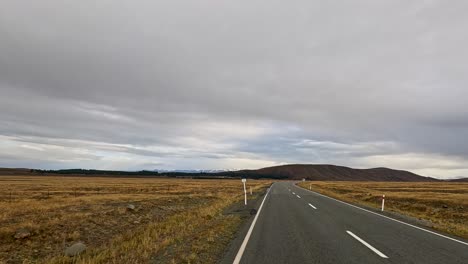 a white campervan travels along an empty rural road through golden autumn fields under overcast skies near lake tekapo, new zealand. static wide-angle shot