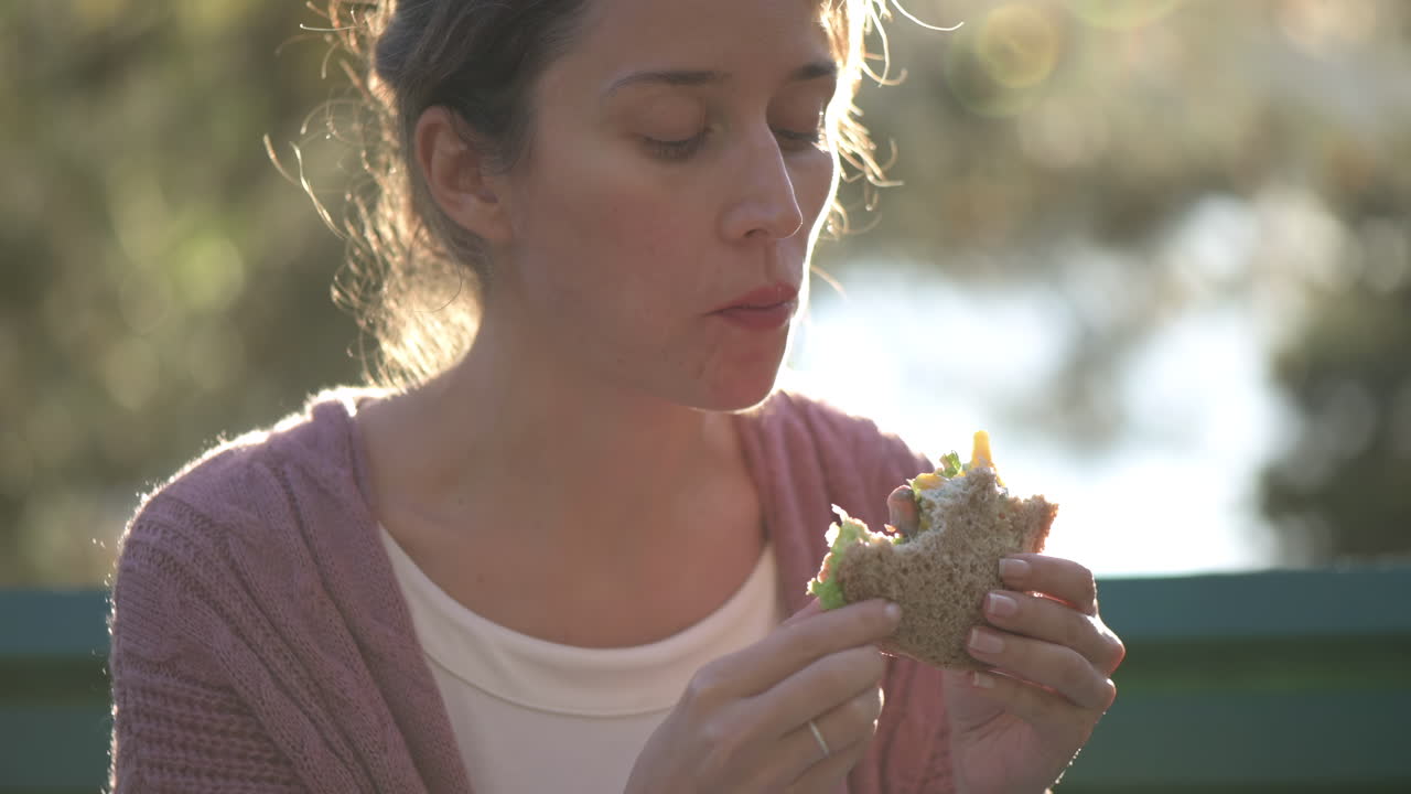 Woman eating a sandwich and writing on paper on a bench in the park