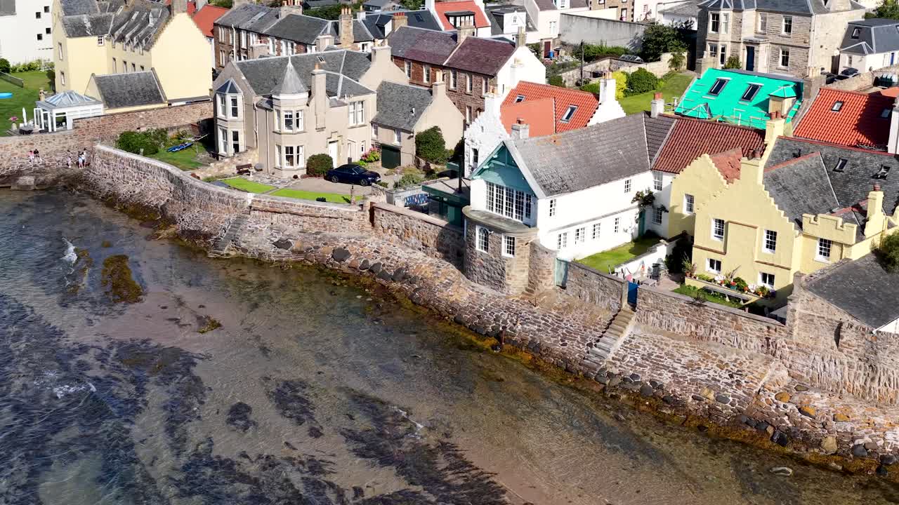 Drone glides above seaside village, historic stone houses, bright daylight, clear sky, tranquil mood