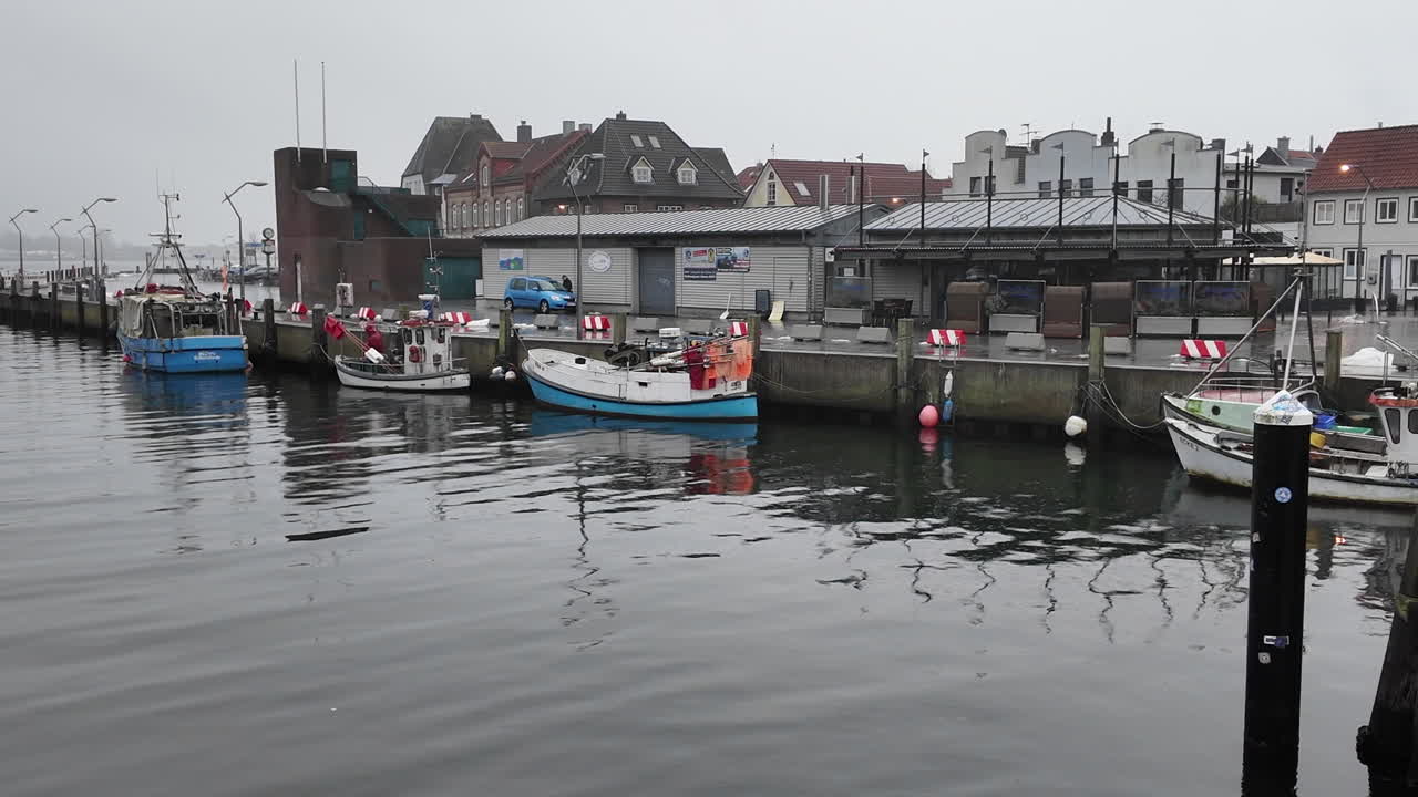 Eine ruhige Hafenszene in Eckernförde, Deutschland, die kleine Fischerboote zeigt, die an einem grauen, bewölkten Tag entlang der Uferpromenade vertäut sind. Traditionelle Hafengebäude, Docks und Spiegelungen auf dem Wasser