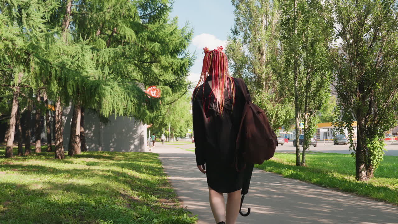 여자가 우산을 쓰고 평온하게 산책하는 모습, 우산을 든 차분한 여성의 모습이 공원을 천천히 거닐고, 화려한 드레드락을 한 상냥한 여자가 햇빛이 비치는 공원의 나무 아래를 천천히 걷고 있어요.