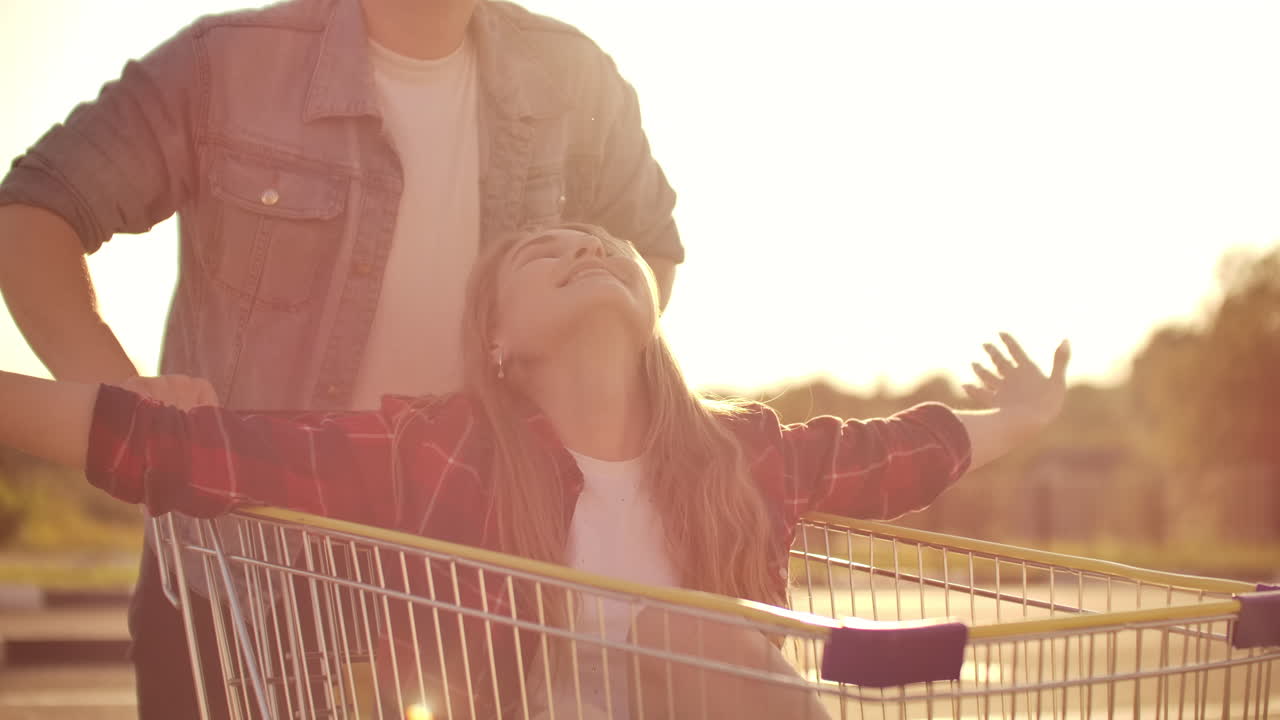 una pareja montando con un carrito de compras en el estacionamiento al aire libre. un joven y elegante cupé divirtiéndose montando con el carrito de tiendas en el estacionamento al aire libre cerca del supermercado
