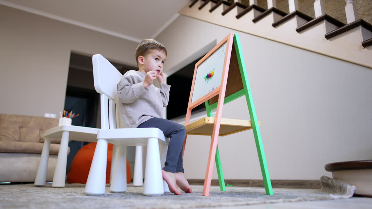 Kid engaging in art at home on a weekend. Young child sits on a chair, focused on drawing at an easel in a cozy living room during a weekend afternoon