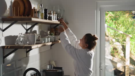 woman takes jar from shelf in kitchen. attractive lady in white gown seeks ingredients for breakfast on shelving unit at home. domestic lifestyle