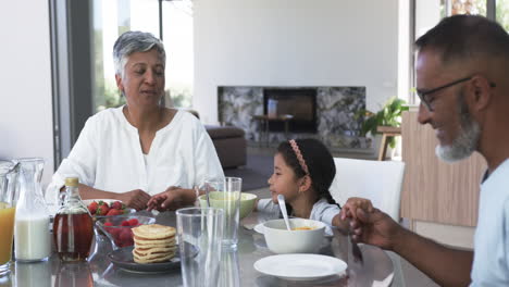 Los-Abuelos-Birraciales-Disfrutan-Del-Desayuno-Con-Su-Joven-Nieta,-Rodeados-De-Panqueques-Y-Frutas.