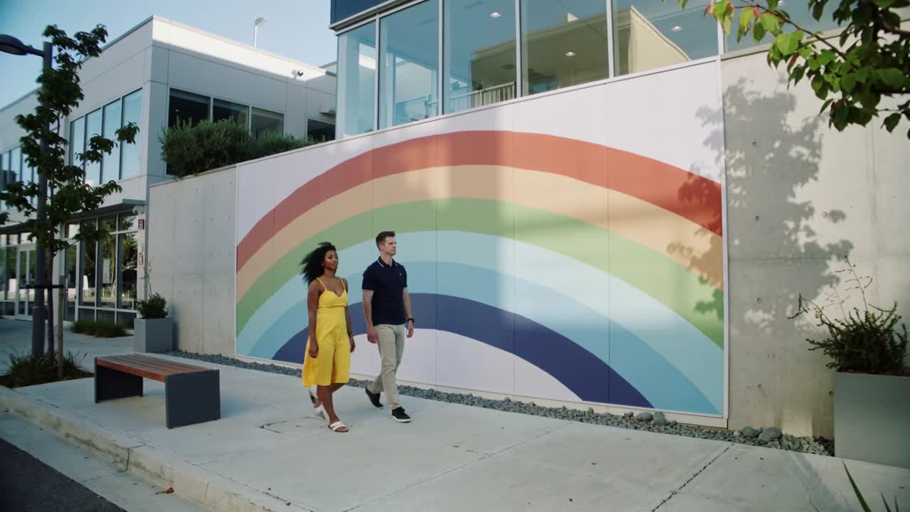 Aproximando-se pela esquerda, casal caminhando por um mural de arco-íris na calçada, mulher de vestido amarelo, homem de polo azul marinho.