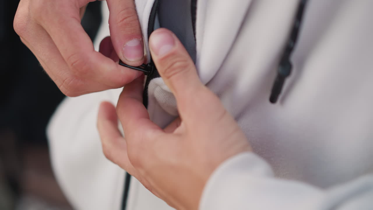 Closeup of zipped hoodie fingers, Person adjusting white hoodie in city rain during quiet evening, Detailed shot of hands fastening zipper on textured jacket with city lights in background