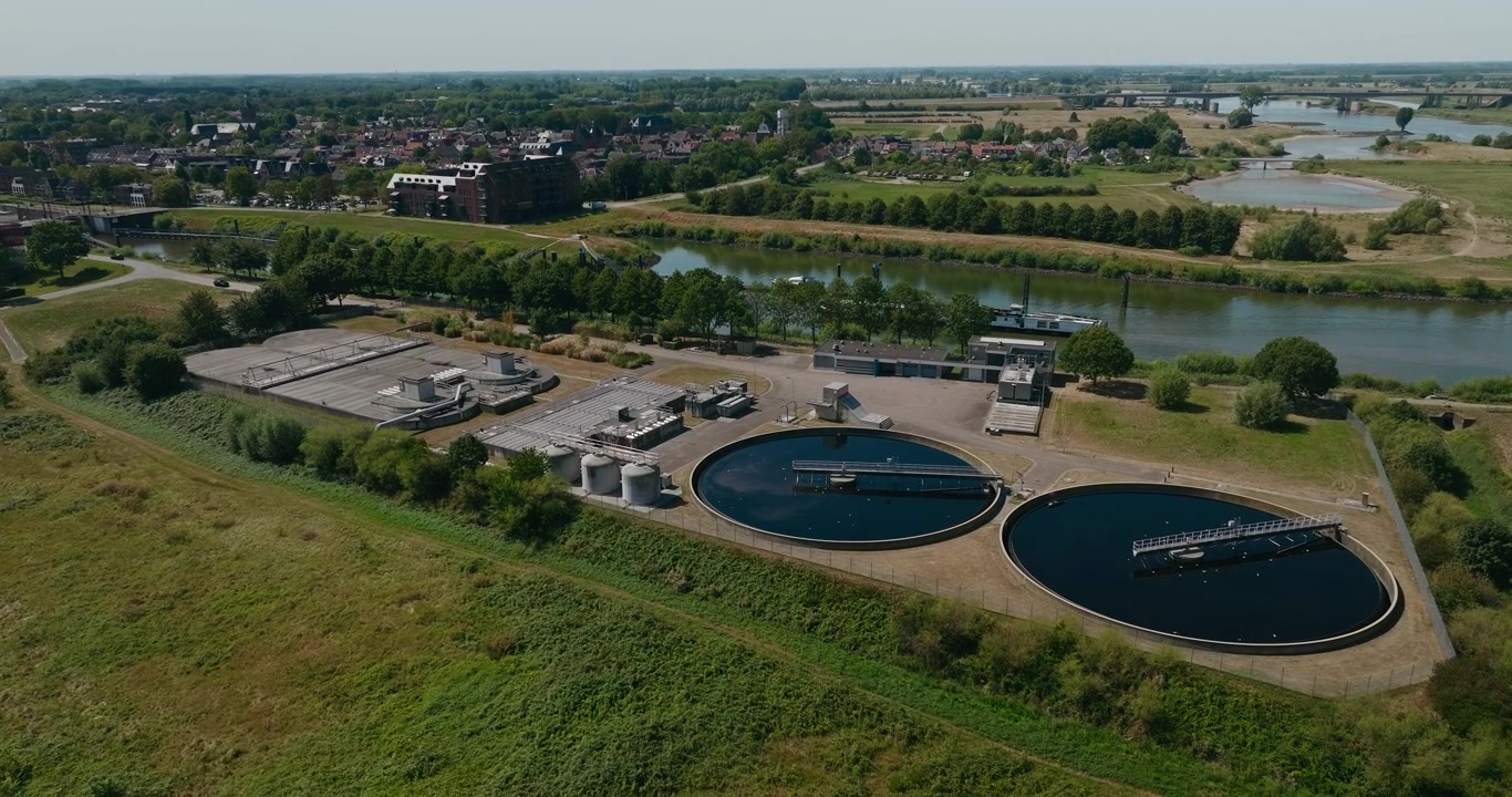 Drone flies toward a wastewater treatment plant with two large basins, while a cargo barge passes on the river behind, connecting water filtration and inland shipping.